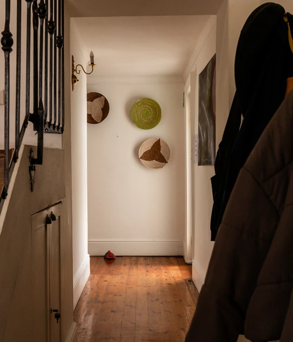 Een zonnige hal met houten vloer, witte muren versierd met drie decoratieve manden, een antieke wandlamp en jassen die aan de rechterkant hangen.